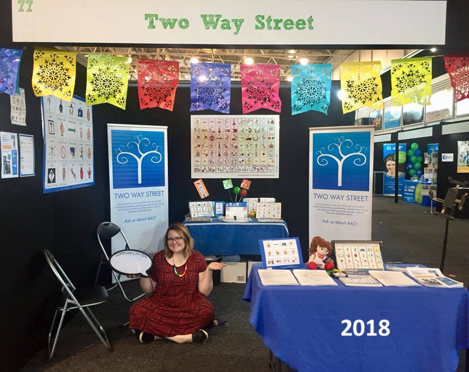 An exhibition booth featuring Two Way Street products and banners, with a Two Way Street staff member sitting on the floor holding a blank speech bubble. The photo is labelled 2018.