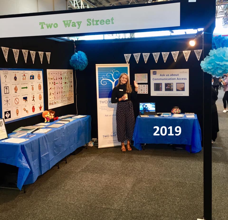 An exhibition booth featuring Two Way Street products and banners, with one Two Way Street staff member, smiling looking at the camera. The photo is labelled 2019.