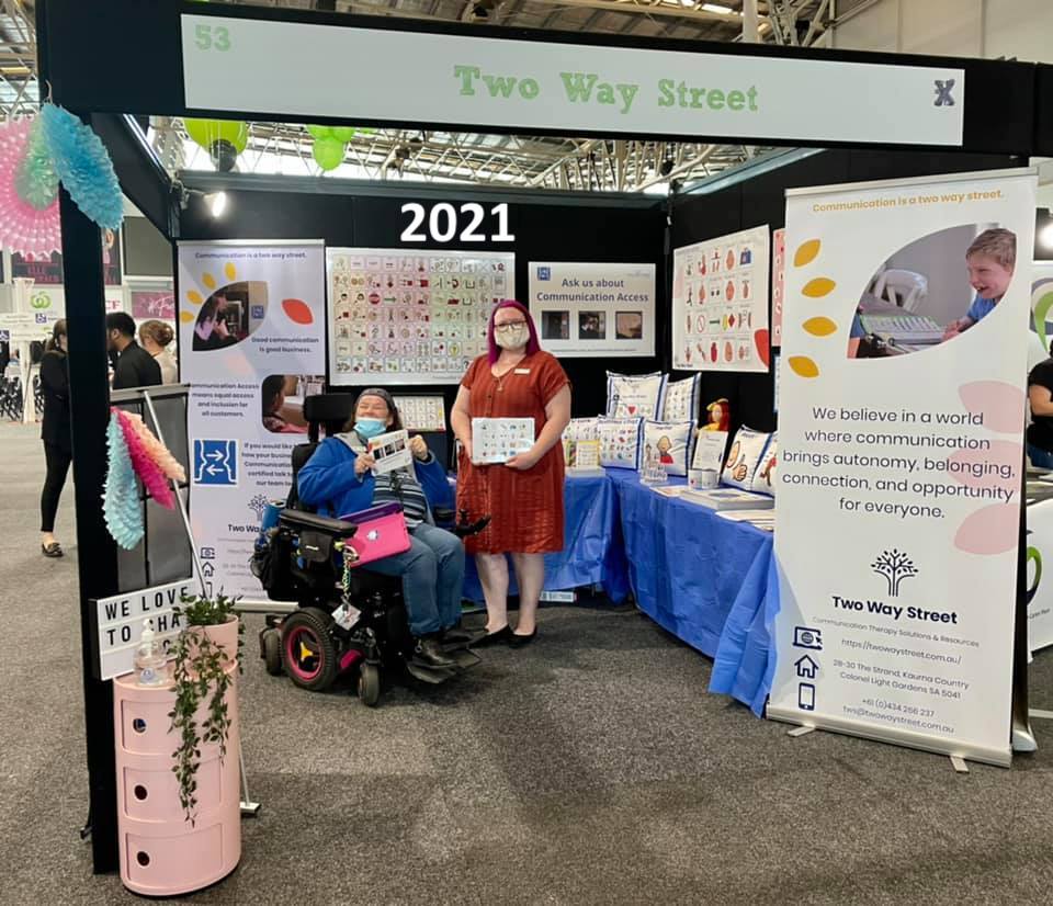 An exhibition booth featuring Two Way Street products and 2 staff members holding communication tools. The photo is labelled 2021.