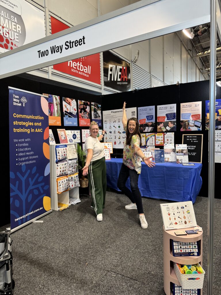 Photo of a colourful Two Way Street exhibition booth with two staff members, arms-wide, showing their product display