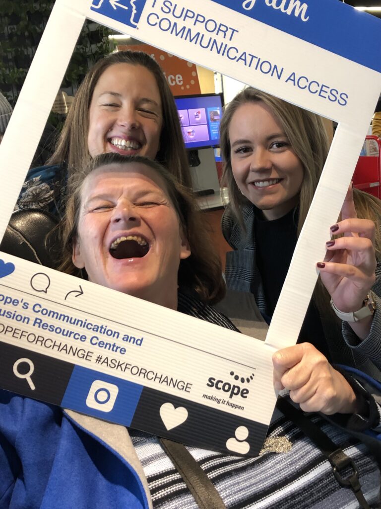 Picture of three smiling Two Way Street staff members, their faces peering through a white cut out frame that says 'I support communication access'.