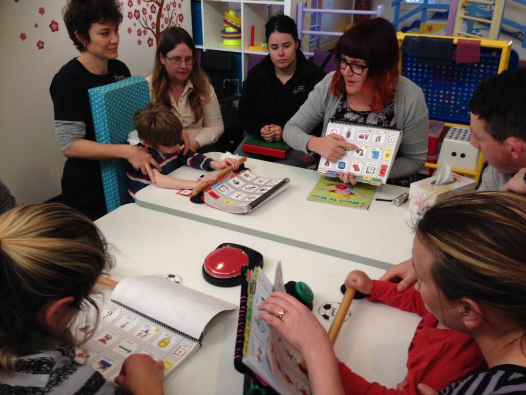 A Two Way STreet staff member holding up and pointing to a symbol in a PODD book, sitting in a classroom with students, teachers and support people, all with their own PODD books