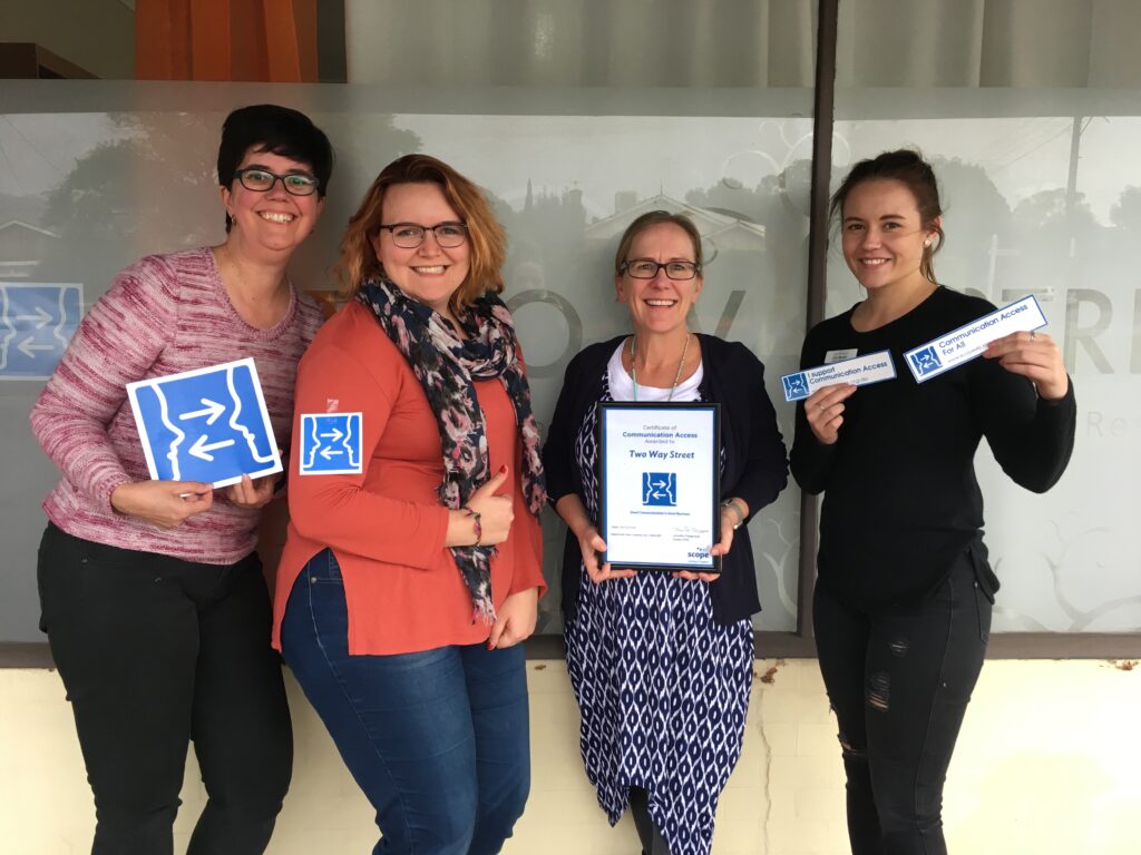 Photo of 4 smiling Two Way Street staff out the front of the Two Way Street office all holding various forms of the communication access symbol and accreditation documentation