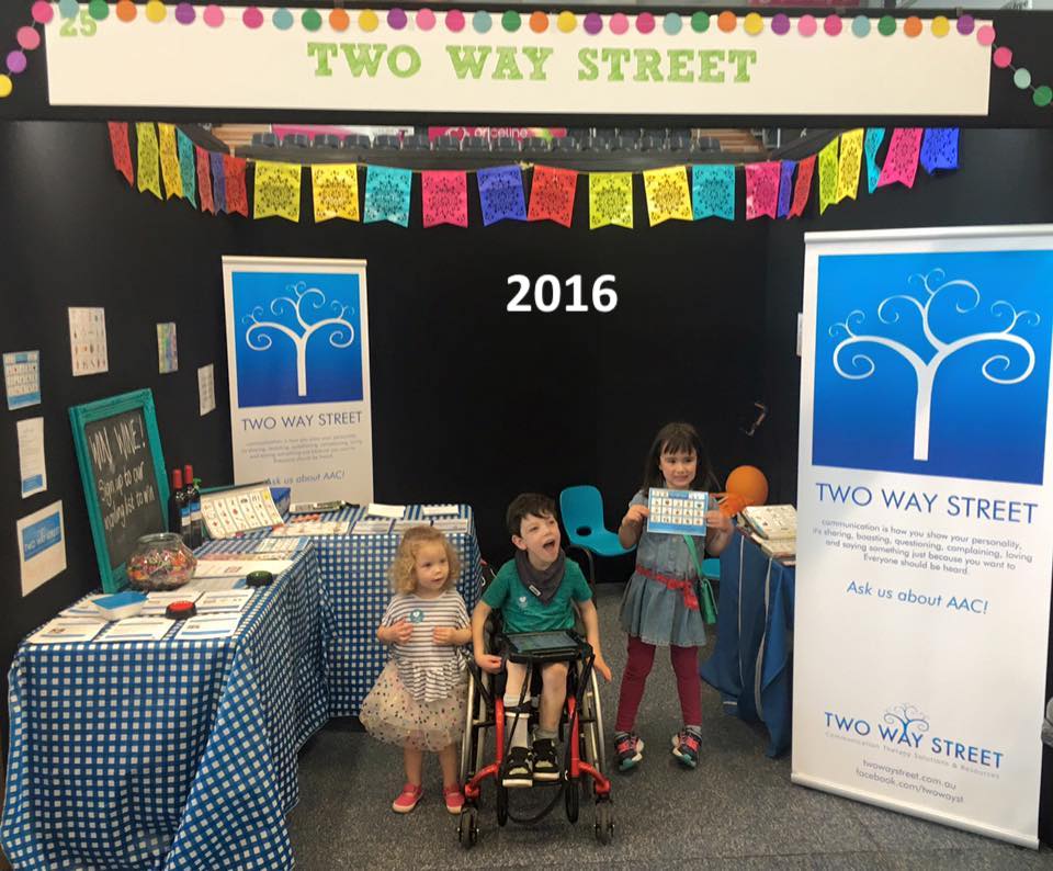 An exhibition booth featuring Two Way Street products and banners, with three children, one in a wheelchair, and one holding a communication tool. The photo is labelled 2016.