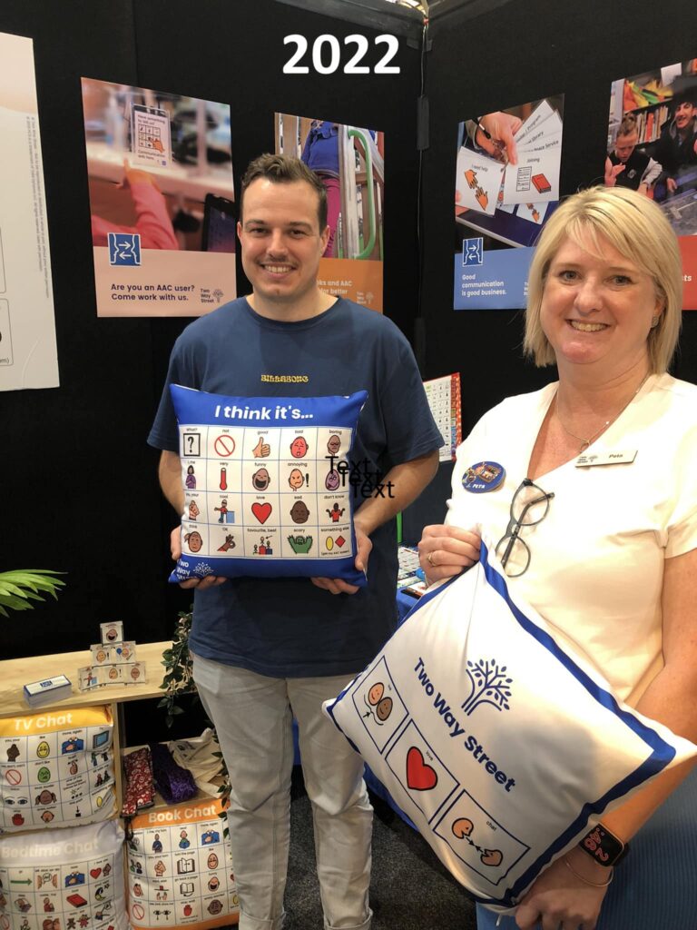 An exhibition booth featuring Two Way Street products and posters, with 2 staff members holding AAC chat cushions The photo is labelled 2022.